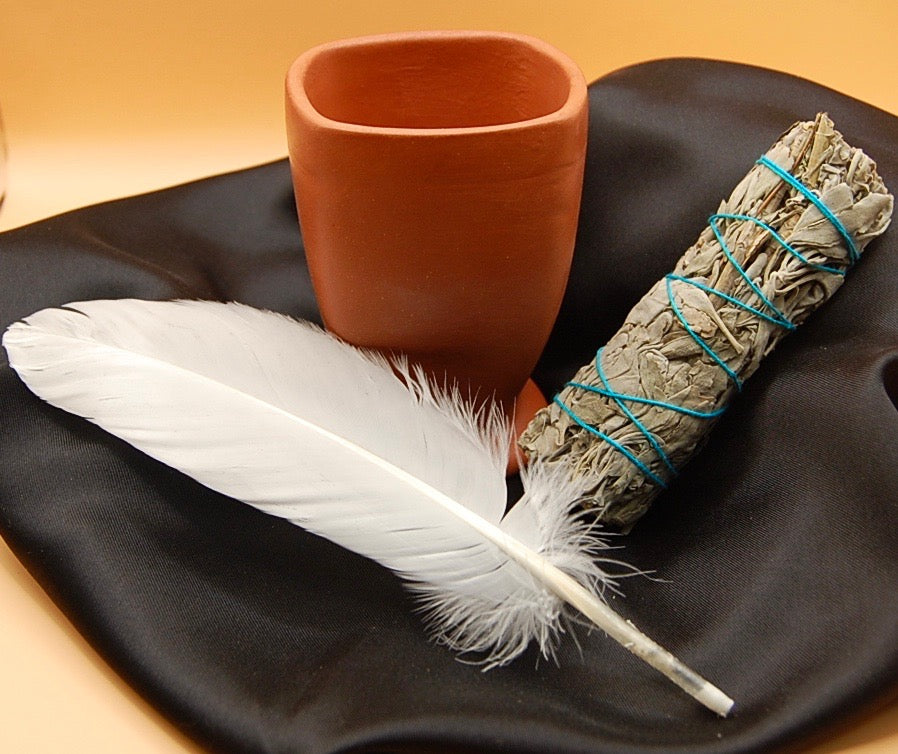 Smudging kit consisting of an organic sage stick, feather and a clay pot on a black napkin